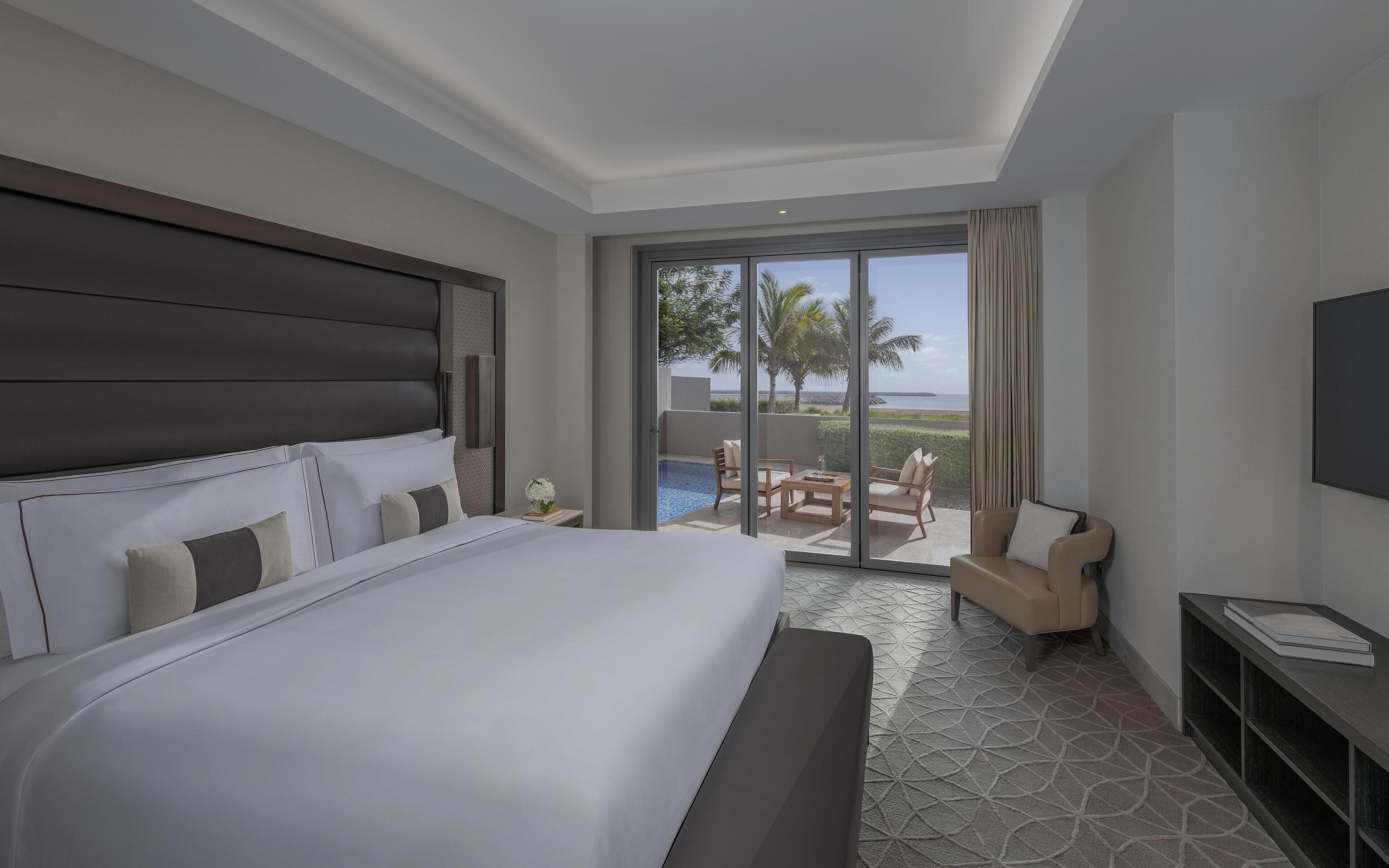 A hotel room with a large bed, a TV, a small table with a book, and a view of a pool and palm trees.