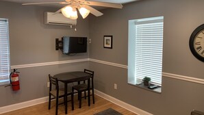 Una habitación con ventilador de techo, televisor de pantalla plana montado en la pared, mesa de comedor con sillas, extintor y ventana con persianas.