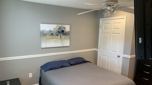 Un dormitorio con cama, ventilador de techo, una puerta y un cuadro en la pared.