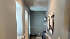 Un pasillo con piso de madera, paredes grises y detalles blancos. Se observa una banca, un cuadro en la pared y una puerta que conduce a otra habitación.