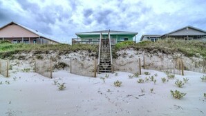 A beachfront property with a wooden staircase leading to a green-roofed building, surrounded by sandy dunes and a wooden fence. - Bright's Beach Front | Sound View + Walk to Beach! (Topsail Beach)