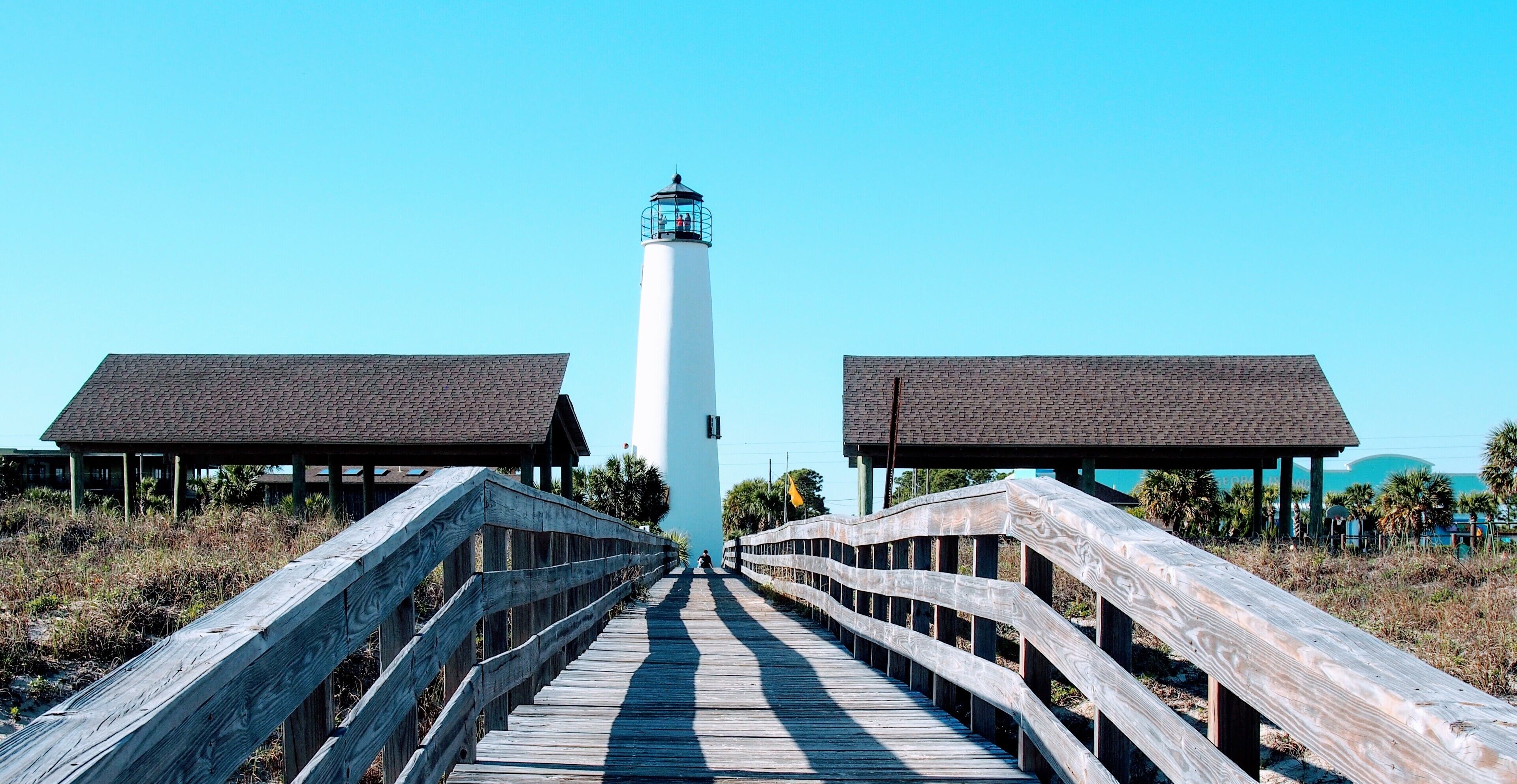 A wooden boardwalk leading to a white lighthouse with a brick roof structure on either side.