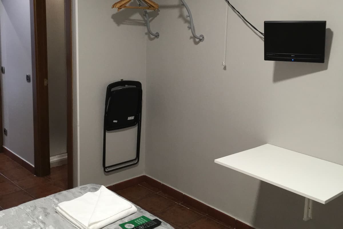 A bedroom with a bed, a wall-mounted TV, a fan, a drying rack, and a small shelf.