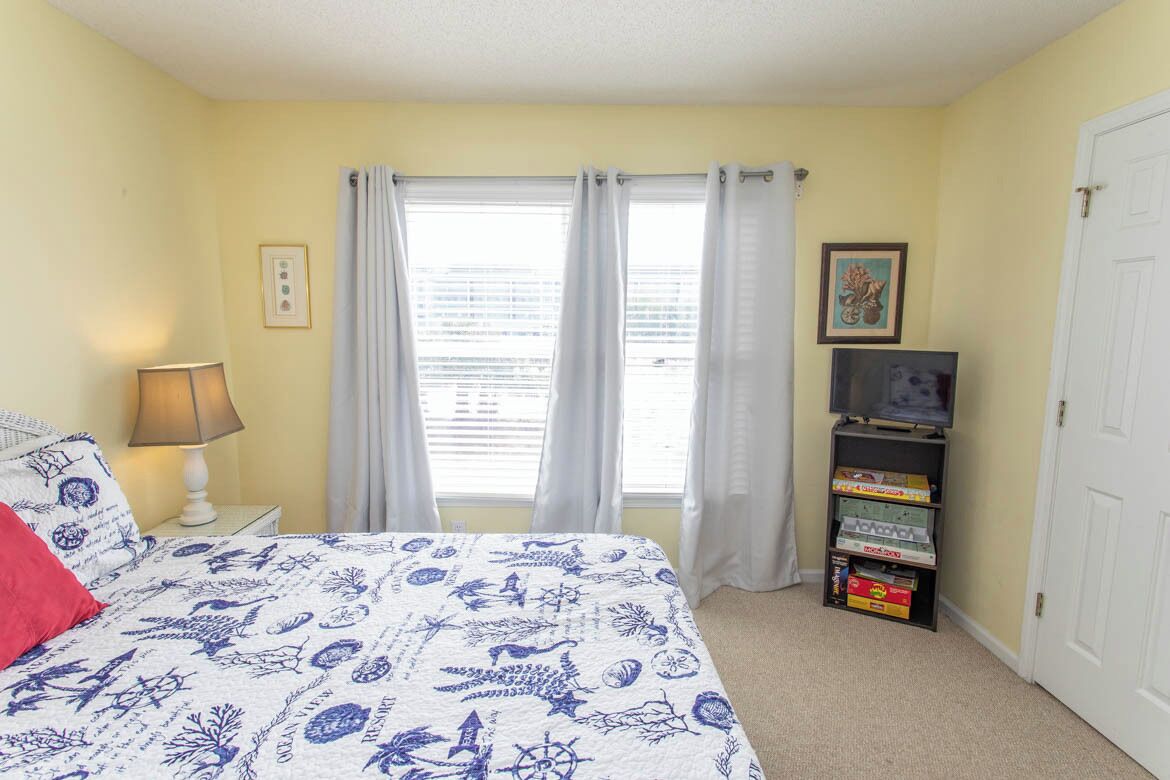 A bedroom with a bed, a nightstand, a lamp, a window with curtains, a television on a stand, and a door.