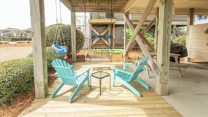 Un espacio de estar al aire libre con techo, con sillas de madera y una mesa pequeña, rodeado de vigas de madera y con vista a un edificio al fondo.