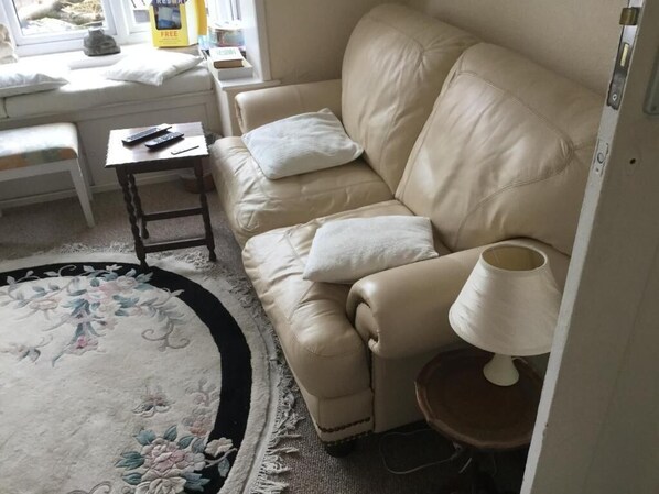 A beige leather sofa with two white cushions, a small wooden side table with a remote control, a lamp on a wooden stool, and a floral rug. - Lakeside Guest House (Weymouth)