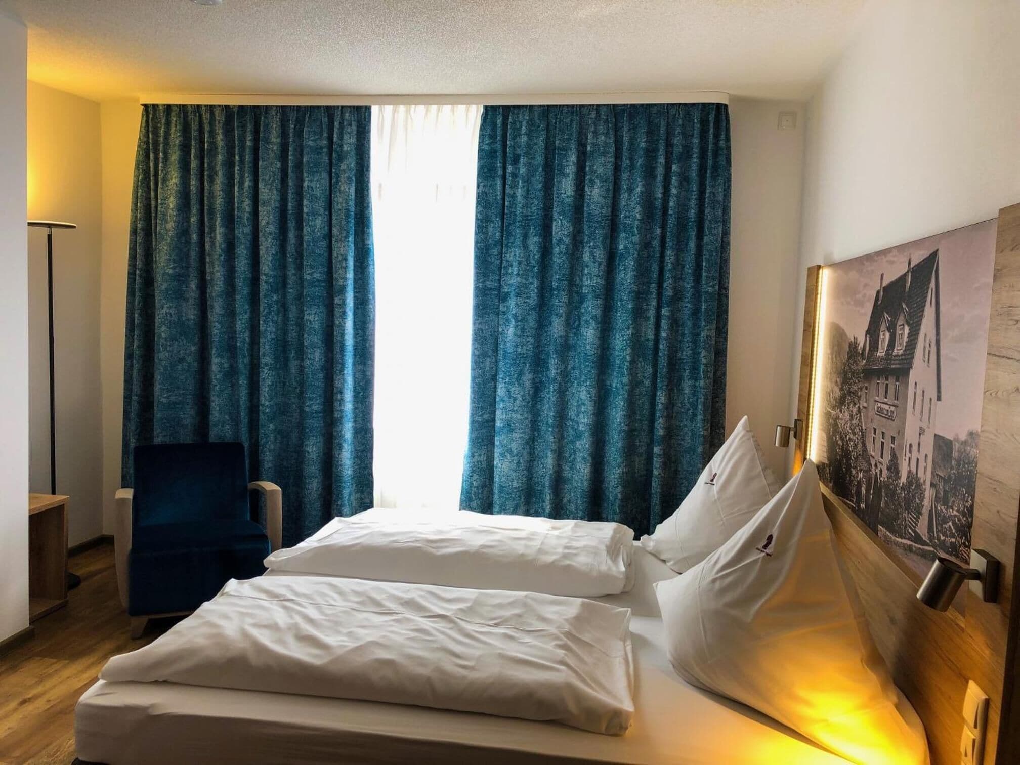 Quarto de hotel com cama, duas almofadas, uma cadeira e uma janela grande com cortinas.