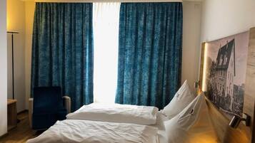 Quarto de hotel com cama, duas almofadas, uma cadeira e uma janela grande com cortinas.