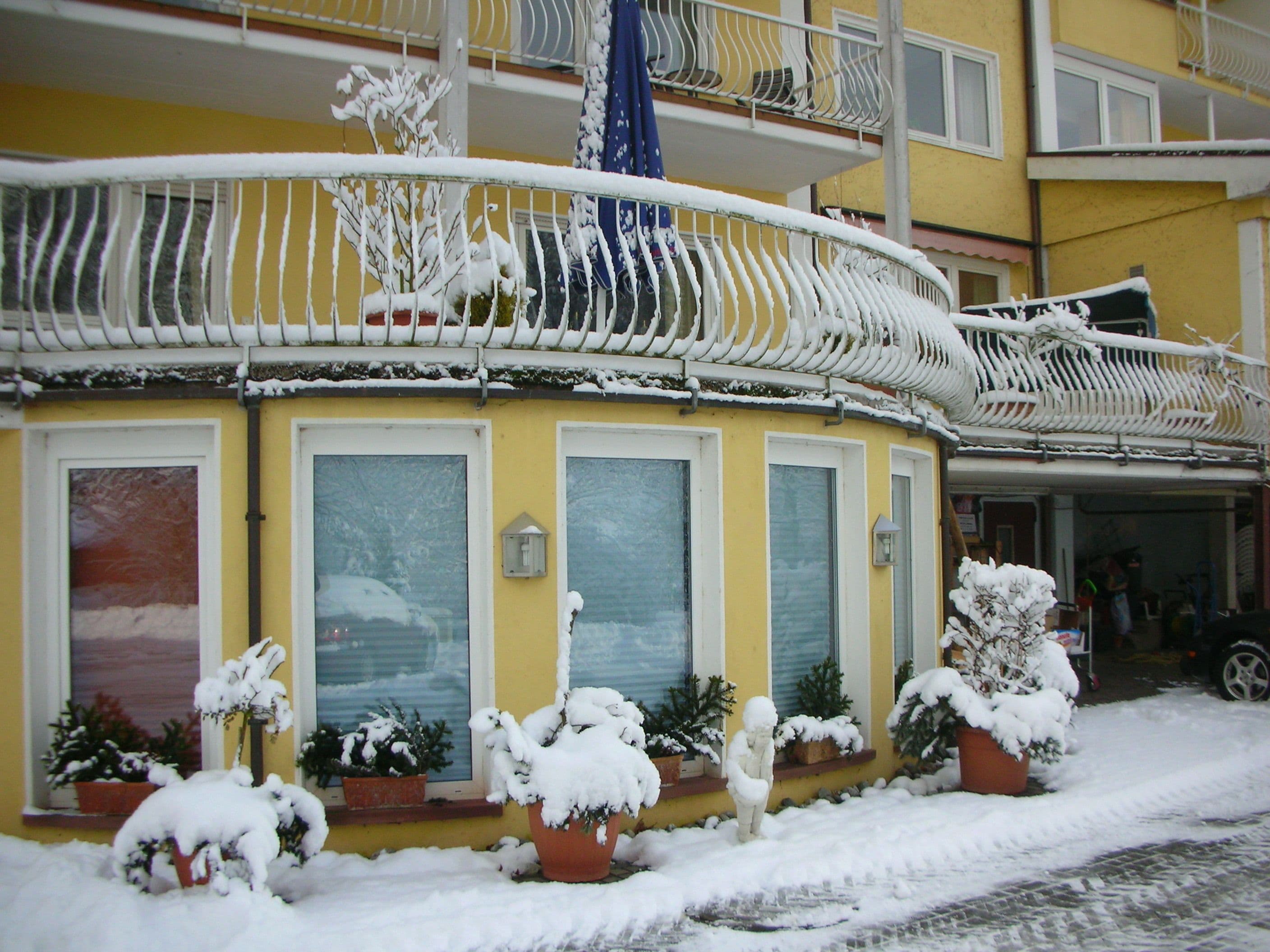 A yellow building with a balcony and potted plants covered in snow.