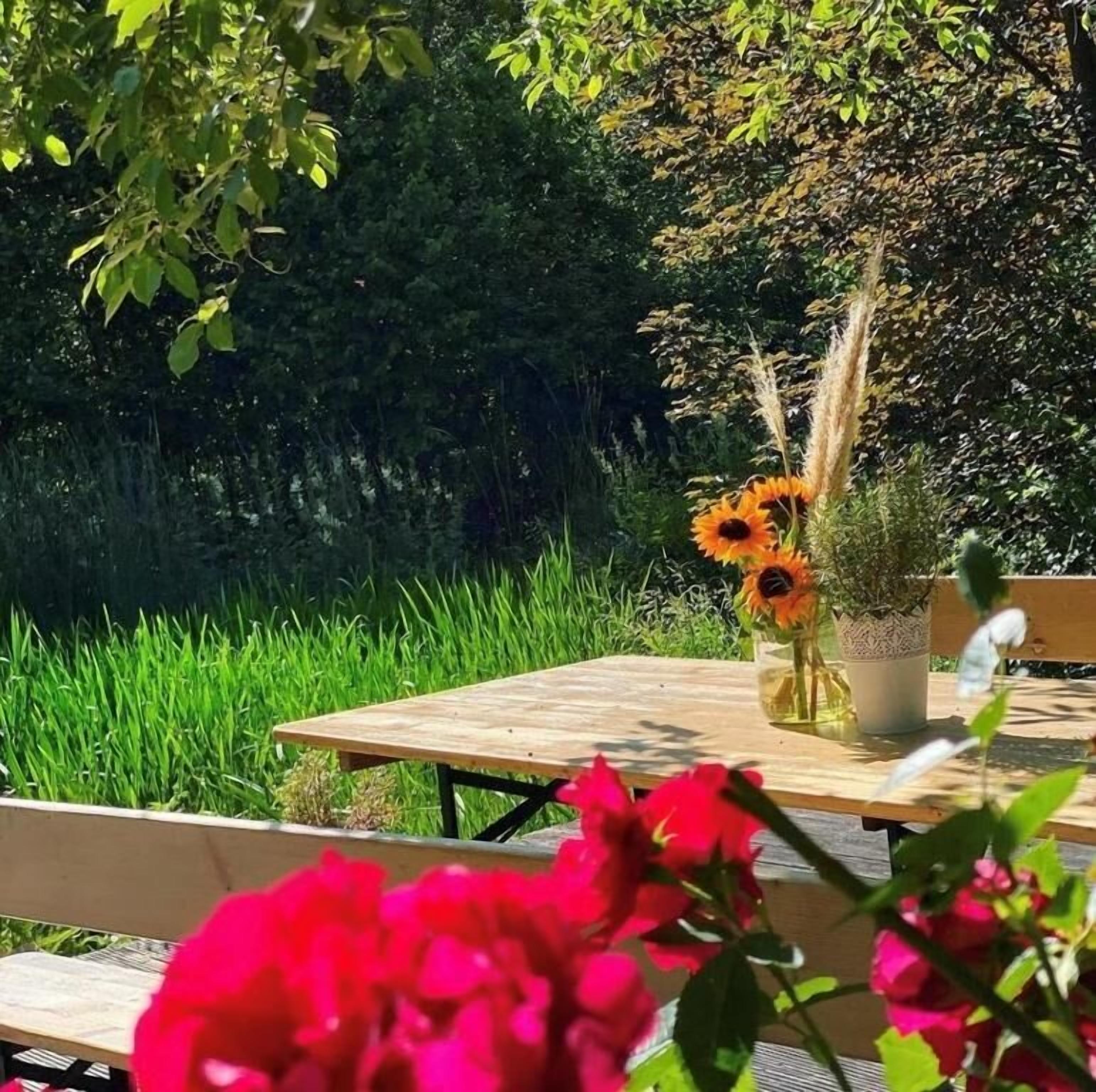 Ein Holztisch mit einer Sonnenblumenvase und einer Topfpflanze, umgeben von Grünpflanzen und einer Bank.