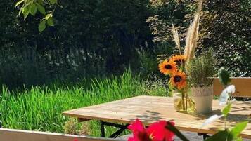 Ein Holztisch mit einer Sonnenblumenvase und einer Topfpflanze, umgeben von Grünpflanzen und einer Bank.