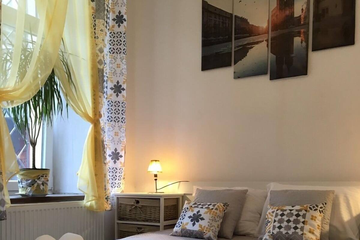 Cama bem arrumada com colcha, almofadas e toalhas dobradas em uma mesa de cabeceira. Há uma luminária na mesinha de apoio e uma janela com cortinas.