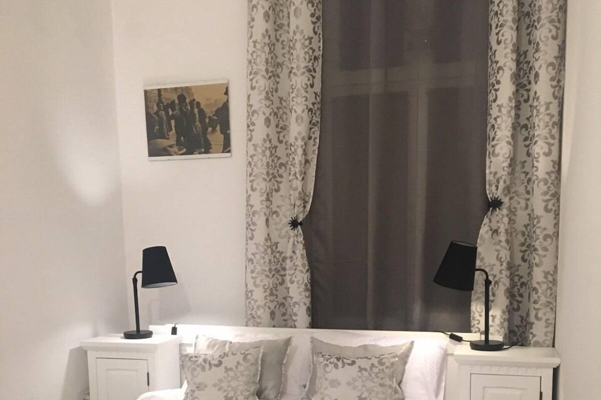 Uma cama bem arrumada com colcha escura e almofadas brancas, duas mesas de cabeceira com luminárias, uma cortina estampada e um pequeno quadro na parede.