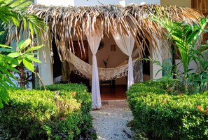 Estructura con techo de paja, una hamaca, rodeada de vegetación cuidada y un sendero de grava.