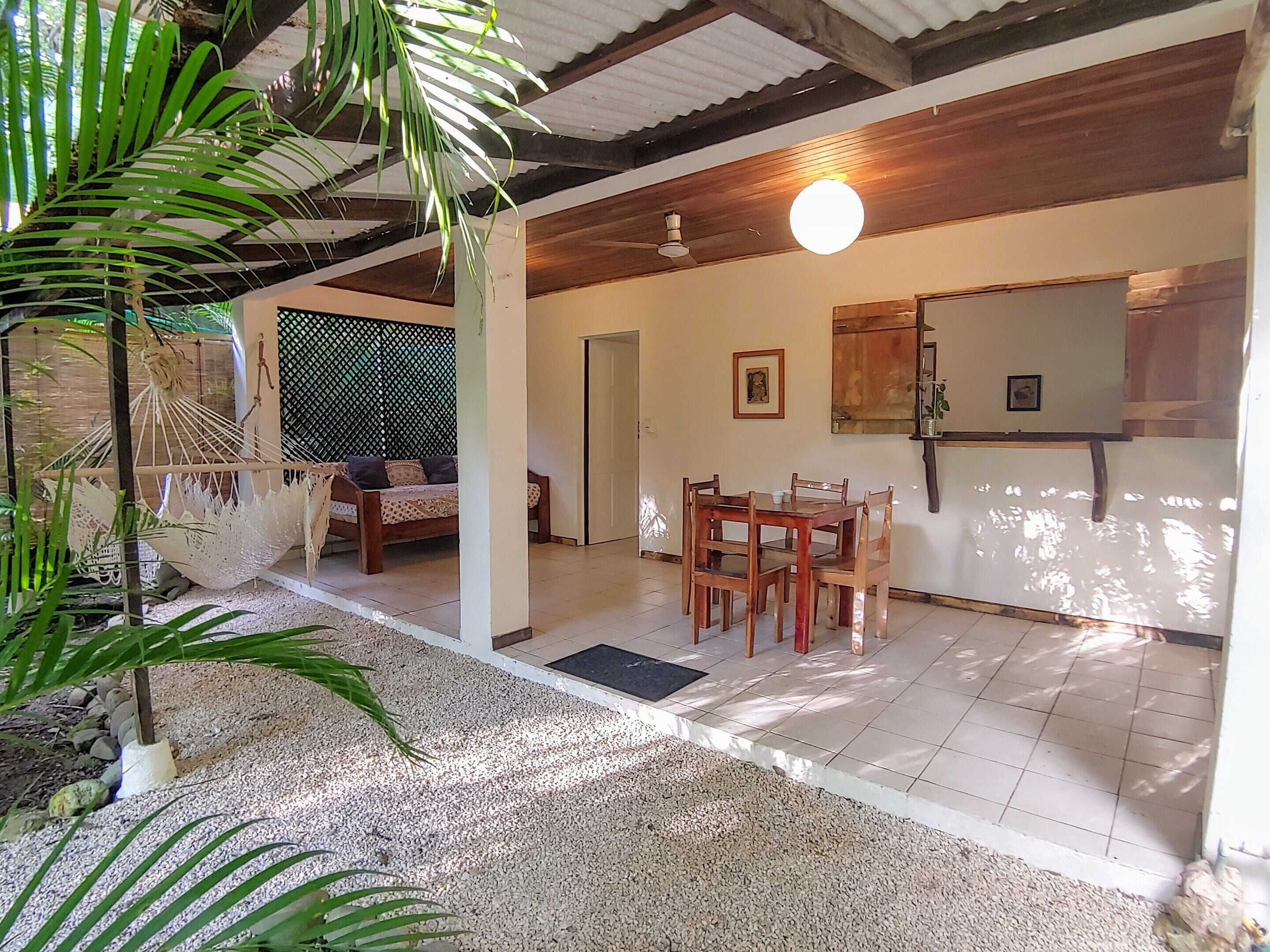 A room with a wooden ceiling, a dining table with chairs, a hammock, and a bed.