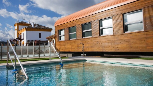 Una piscina con un deck de madera y un edificio con grandes ventanales.