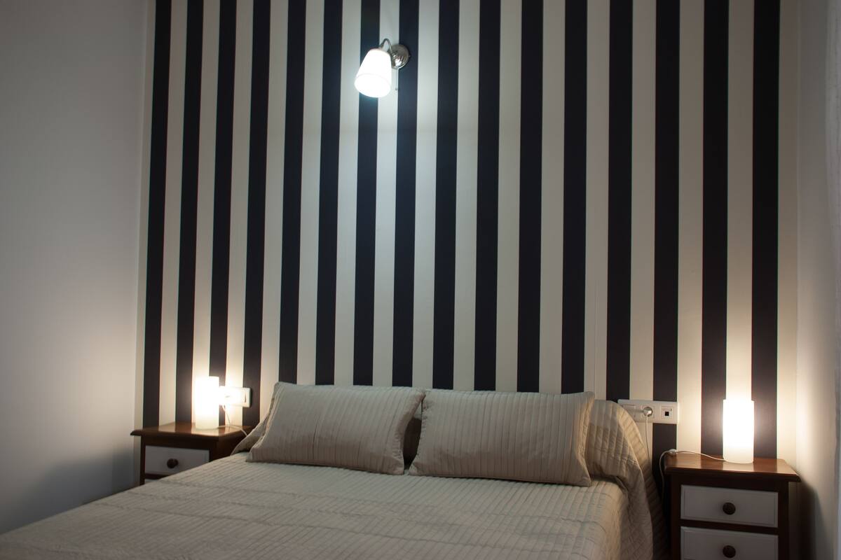 Une chambre avec un mur à rayures, un lit avec deux oreillers et deux tables de chevet avec des lampes.