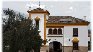 Un edificio de dos pisos con fachada blanca y detalles amarillos, que presenta un arco central y una torre de campana.
