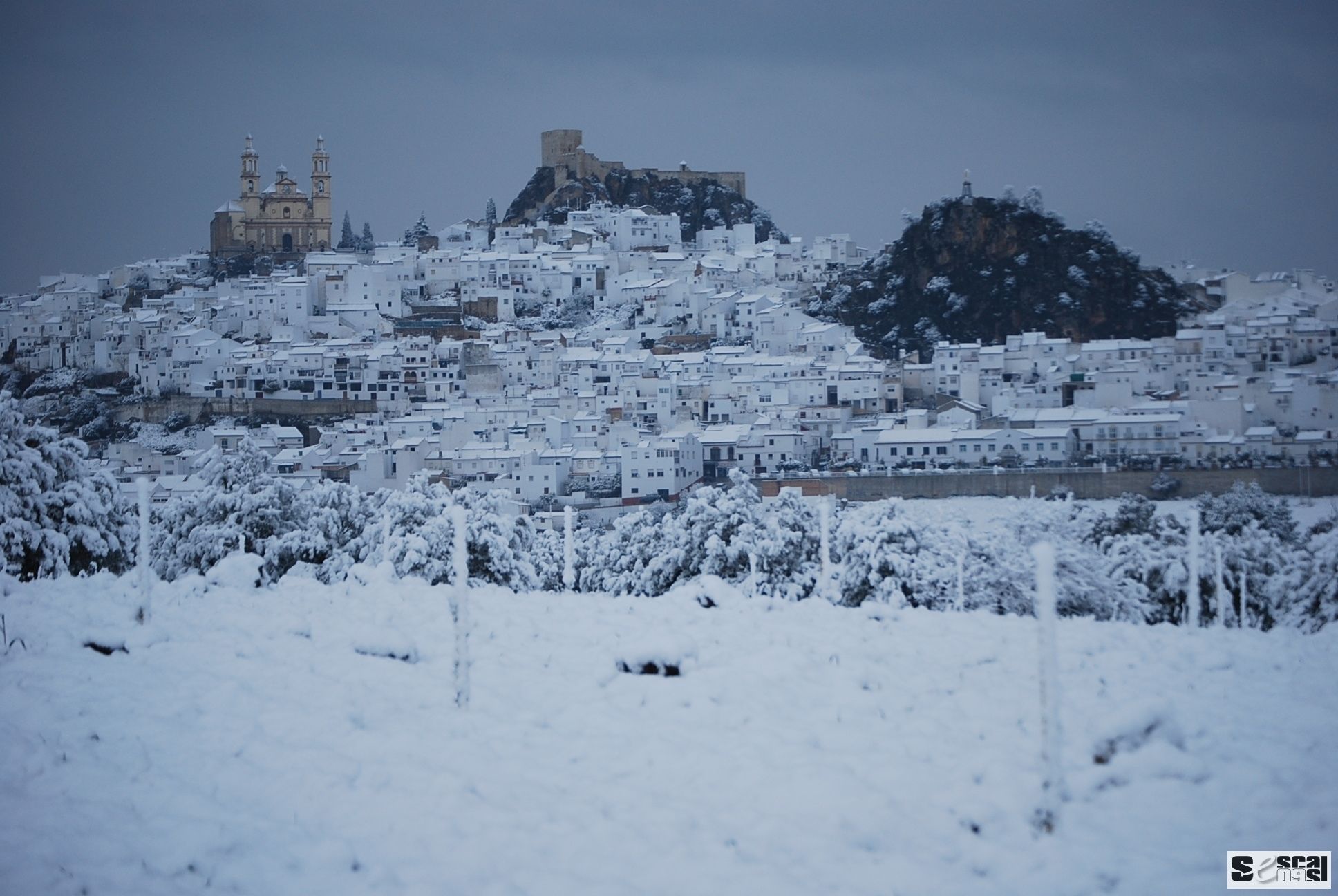 Un pueblo cubierto de nieve con edificios, una iglesia destacada y un castillo en una colina.