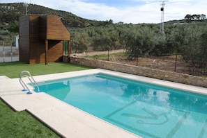 A swimming pool with a wooden structure and a stone wall, surrounded by greenery and hills. - Hostal Vía Verde de la Sierra (Olvera)
