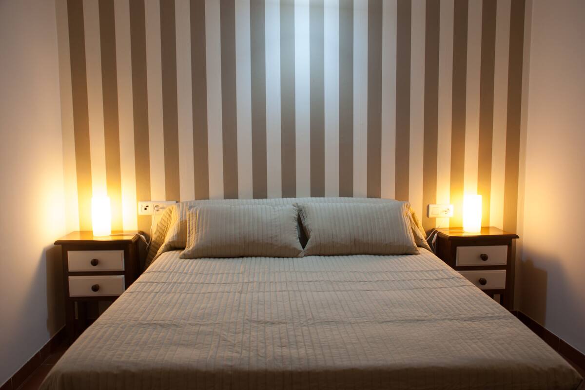 Un lit bien fait, recouvert d’une courtepointe matelassée, deux tables de chevet avec des lampes, et un mur à rayures en arrière-plan.