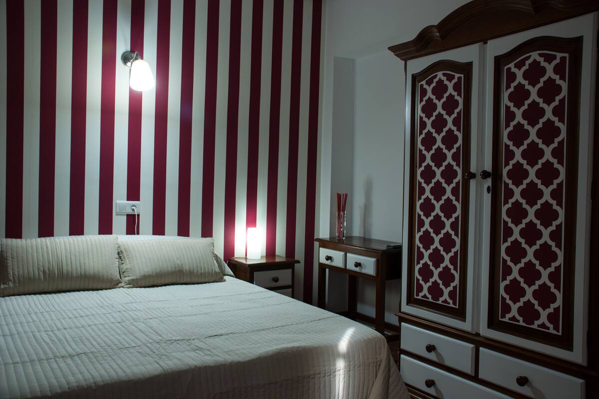Une chambre à coucher comprenant un lit, une table de chevet, une commode aux portes à motifs et un mur orné de rayures verticales.