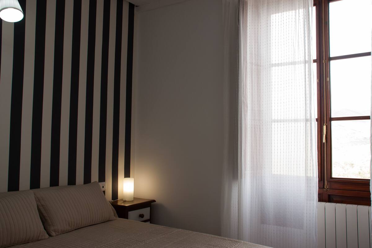 Une chambre avec un papier peint à rayures, une lampe de chevet, une fenêtre avec des rideaux légers et un radiateur.