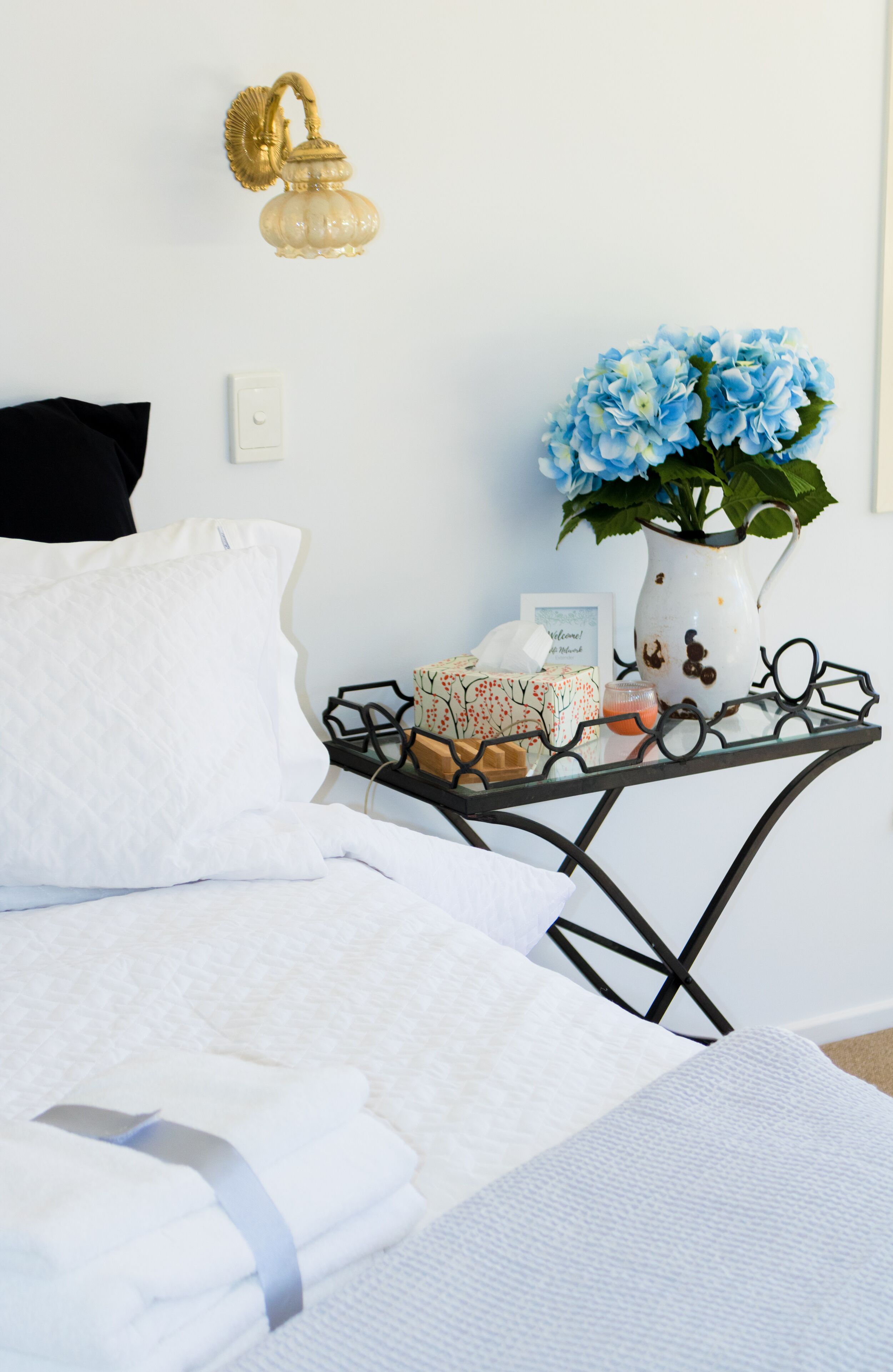 A neatly made bed with white linens, a black pillow, and a white pillow. A small table with a vase of blue flowers, a candle, and a framed picture is beside the bed.