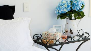 A neatly made bed with white linens, a black pillow, and a white pillow. A small table with a vase of blue flowers, a candle, and a framed picture is beside the bed.