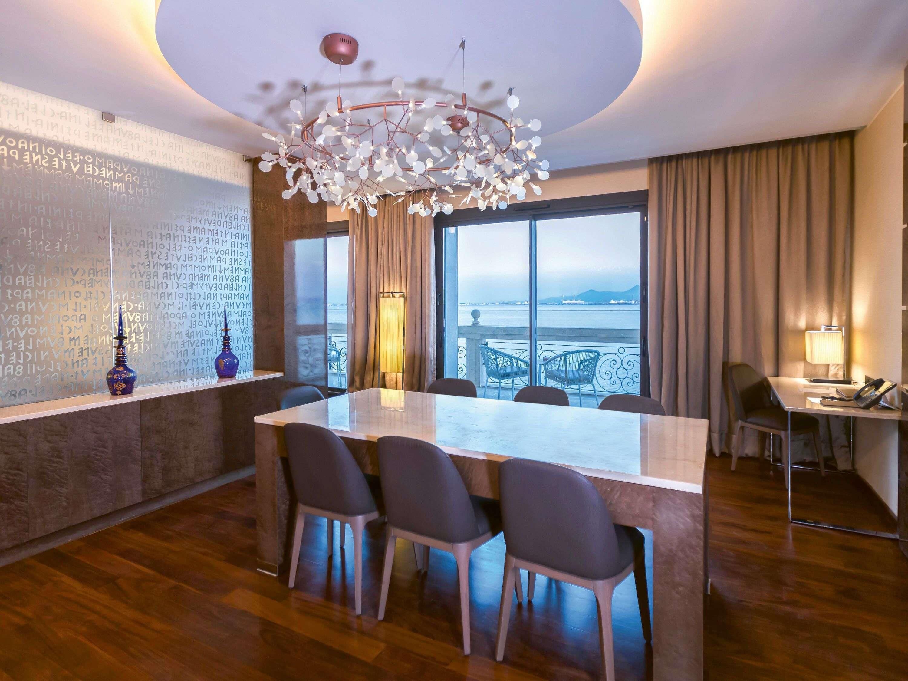 A modern hotel room with a large dining table, four chairs, a chandelier, a large window with a view, and a desk with a lamp.