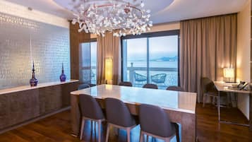 A modern hotel room with a large dining table, four chairs, a chandelier, a large window with a view, and a desk with a lamp.