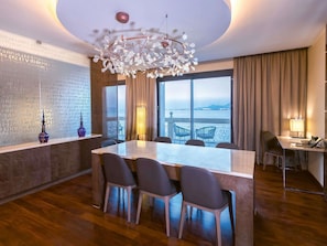 A modern hotel room with a large dining table, four chairs, a chandelier, a large window with a view, and a desk with a lamp. - Mövenpick Hotel du Lac Tunis (Tunis)