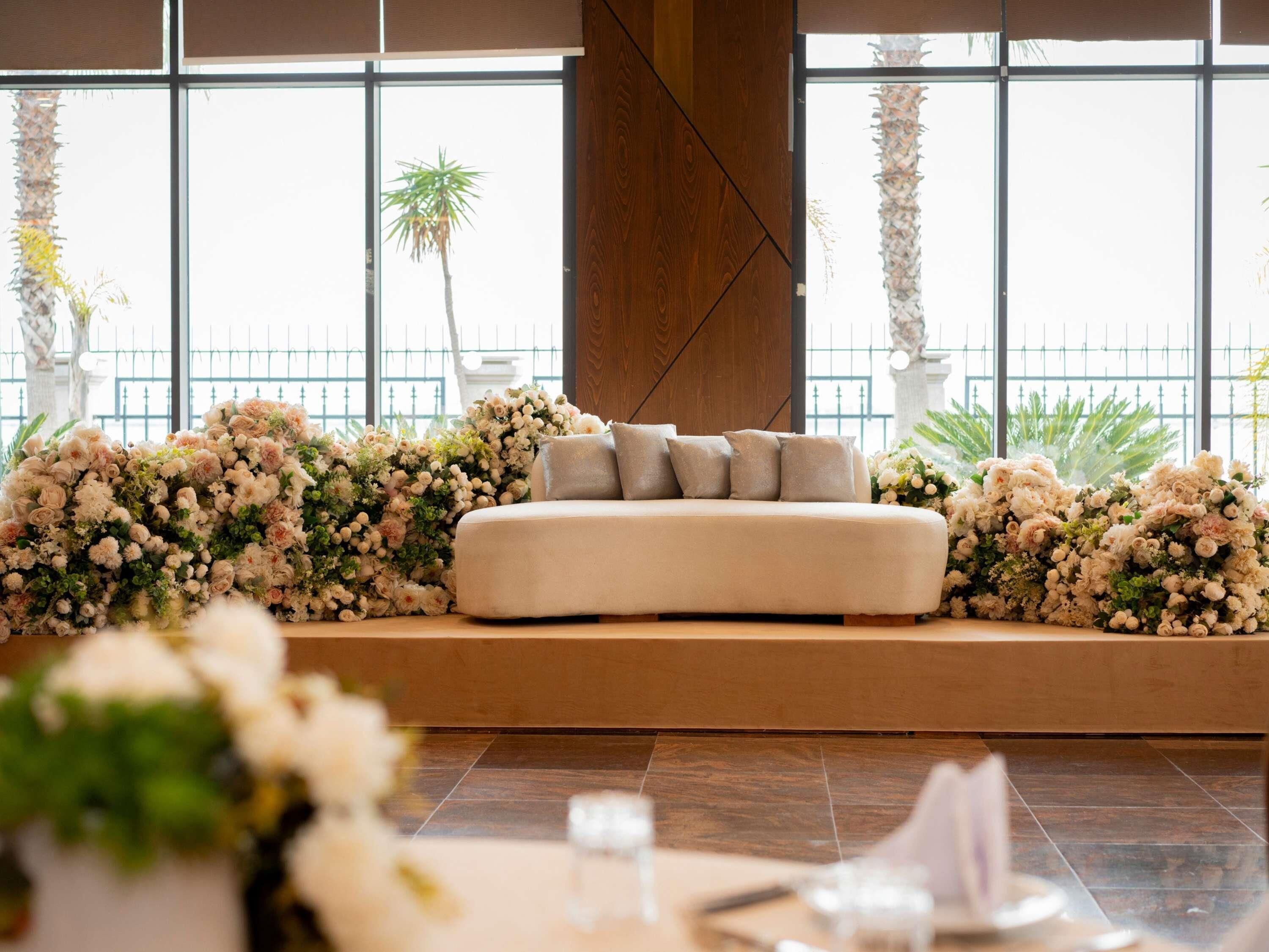 A seating area with a sofa and cushions, surrounded by floral arrangements, in a room with large windows and a view of palm trees.