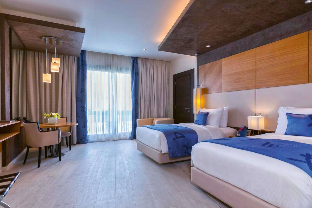 Une chambre d’hôtel avec deux lits, un bureau, une chaise et une grande fenêtre avec des rideaux.