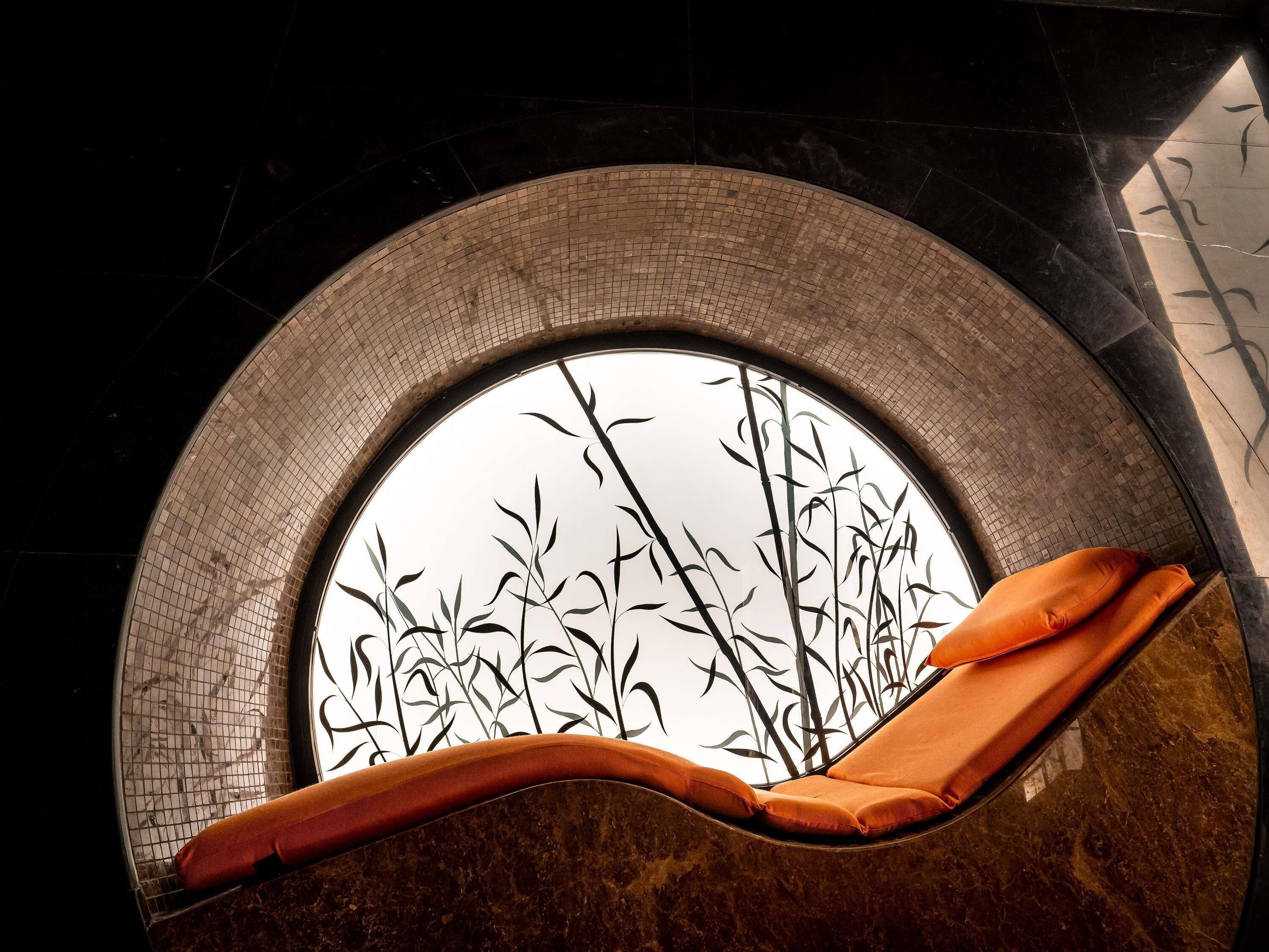 Una ventana circular con diseño de bambú, una silla acolchada y una pared con azulejos.