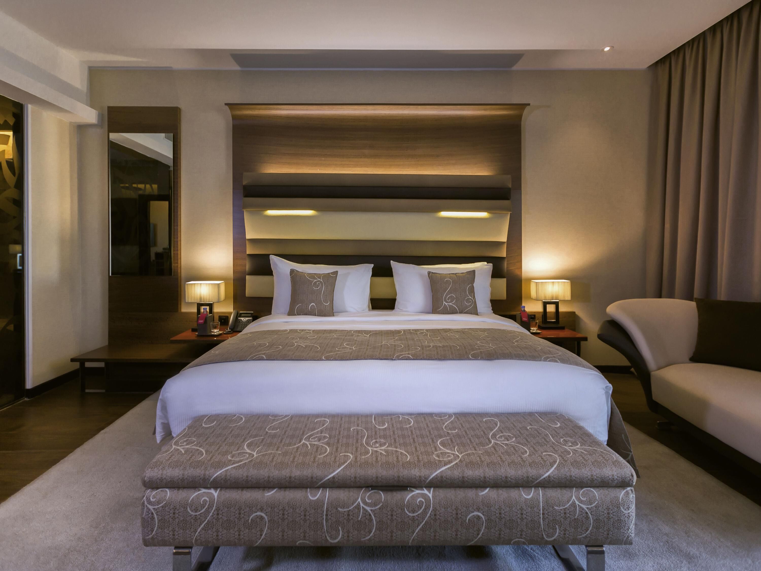 Een moderne hotelkamer met een groot bed, bedlampen, een stoel en uitzicht op een gang.