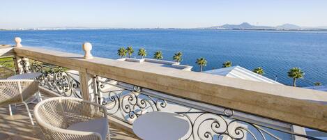 A balcony with wicker chairs and a table overlooking a body of water with palm trees and distant mountains.