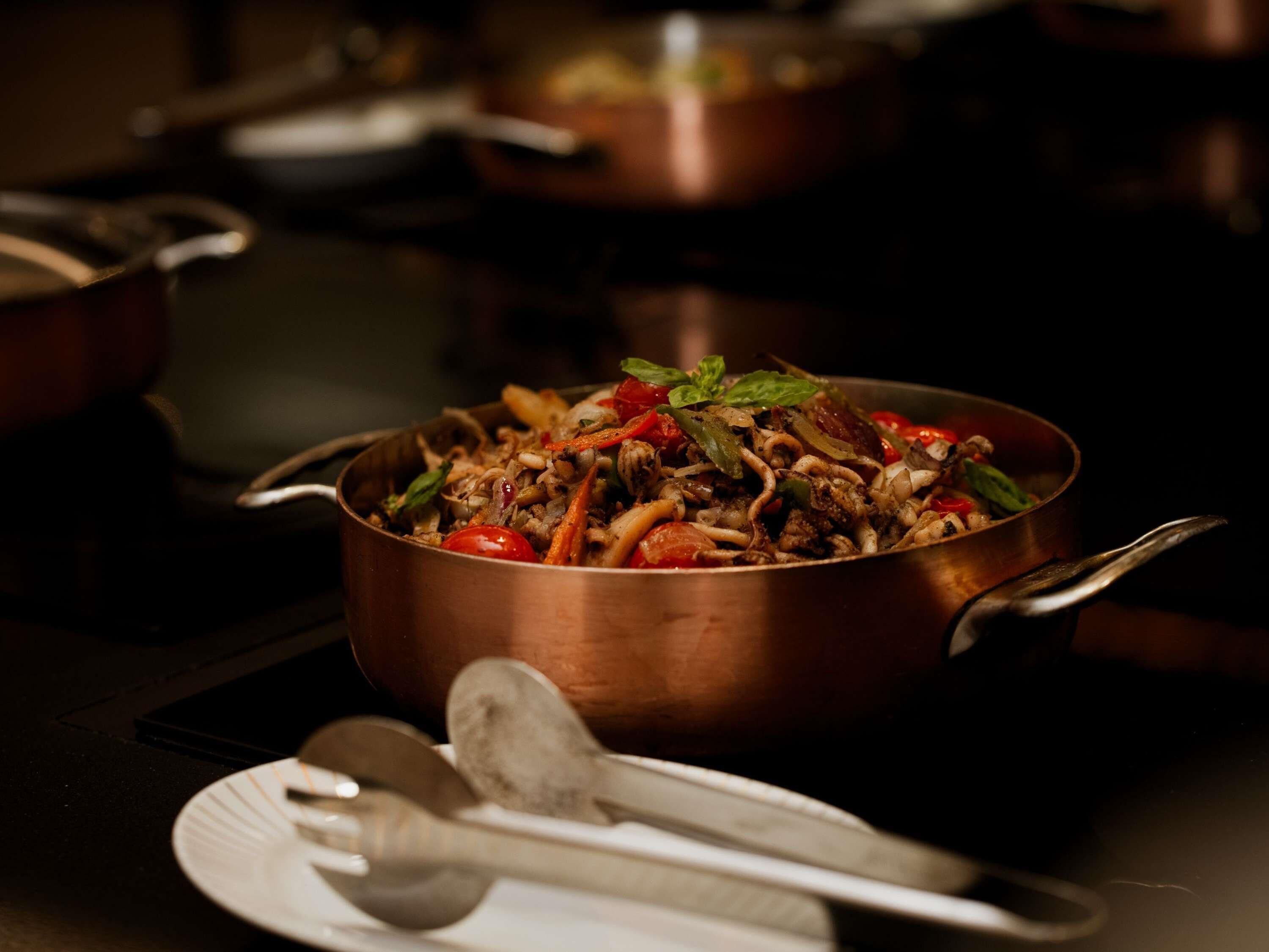 A copper pan filled with a stir-fry dish on a stove, with a spoon and a plate nearby.