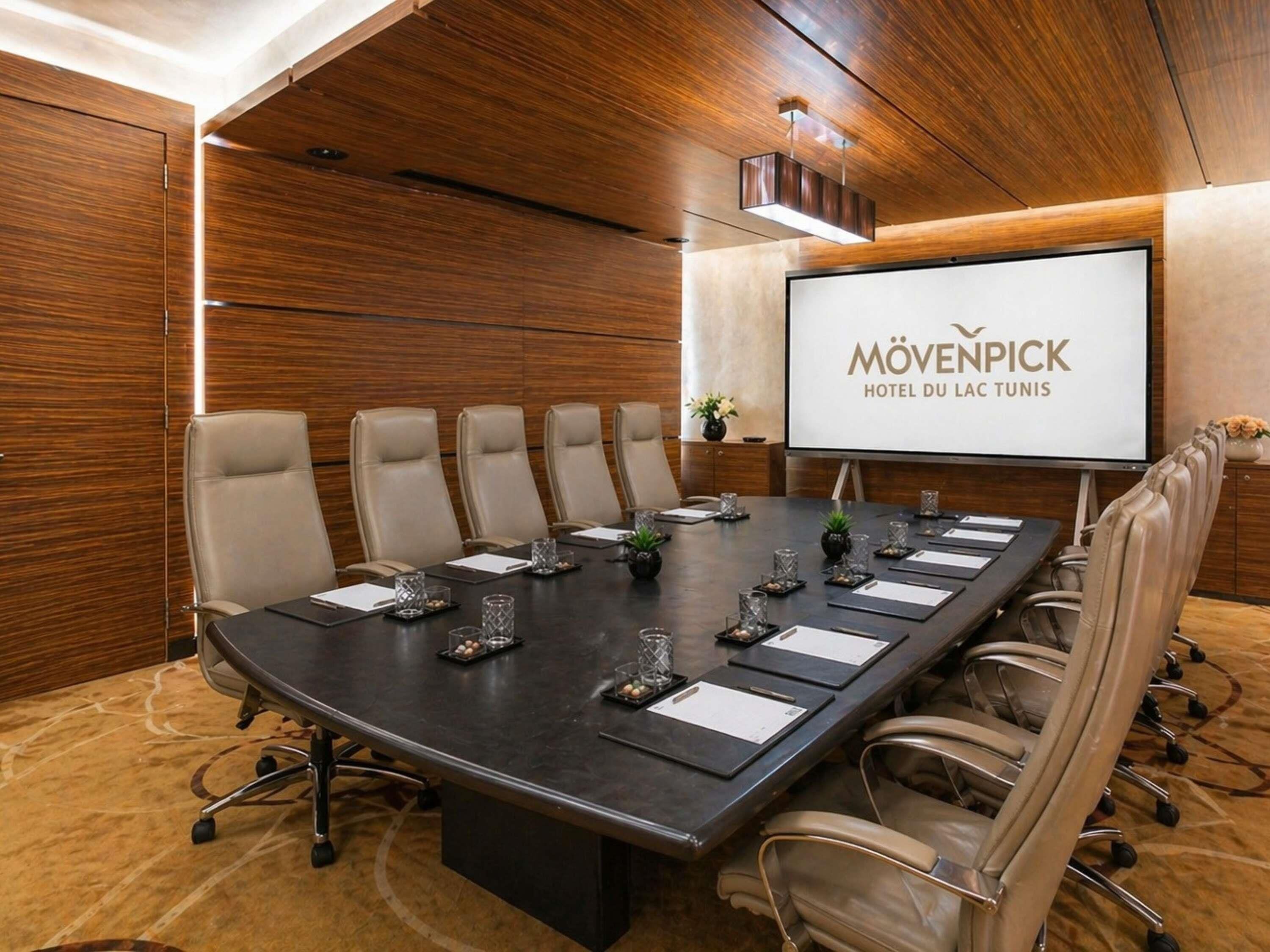 A conference room with a long table, six chairs, a projector screen, and a wooden wall.