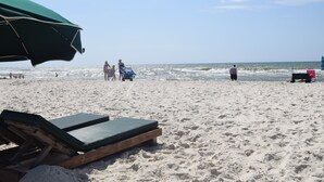 A beach with a lounge chair and an umbrella, a person walking, and a jet ski in the background.