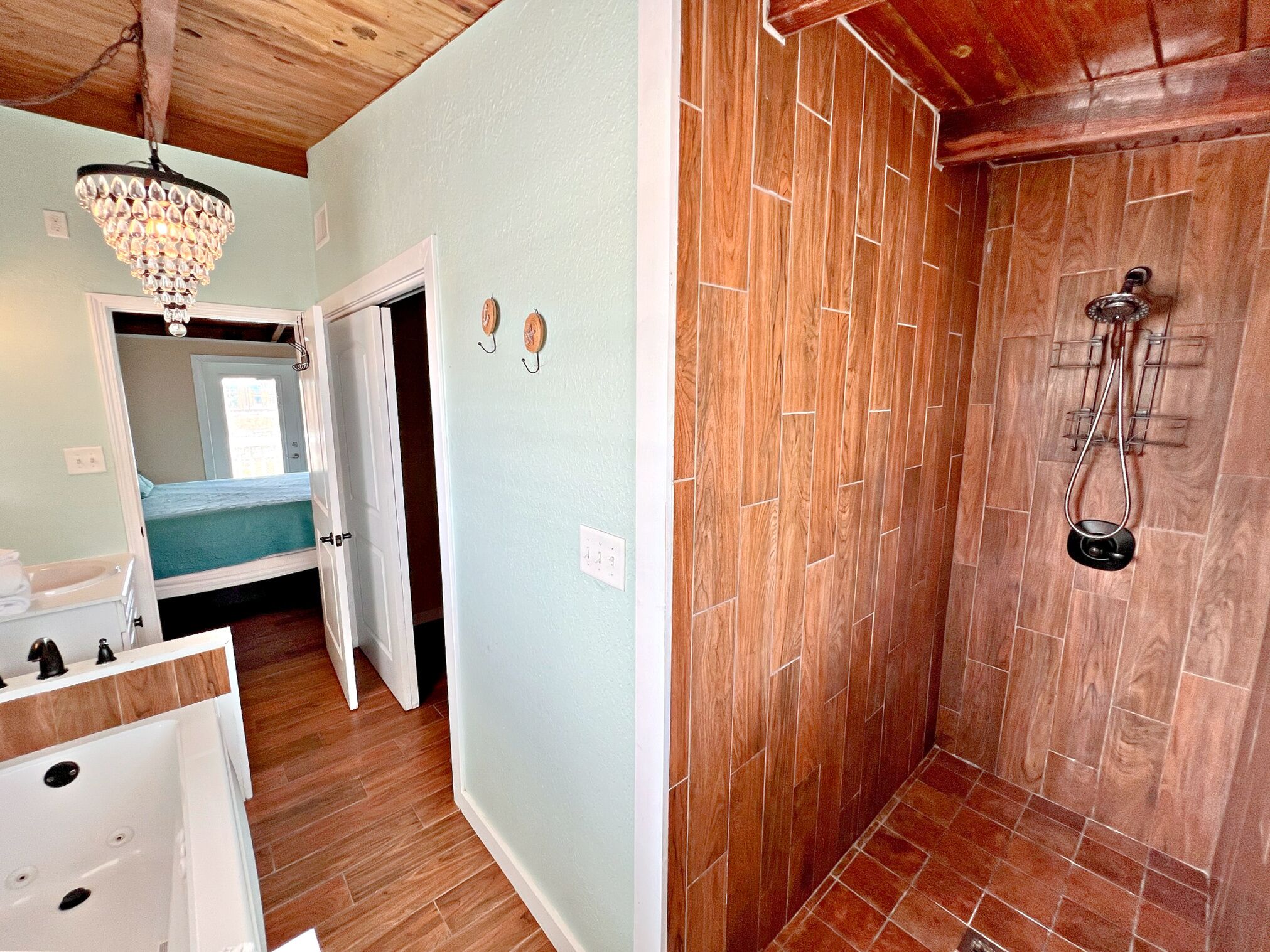 A room with a wooden floor, a white bathtub, a chandelier, and a wooden shower enclosure.
