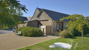 Un bâtiment rustique en pierre, doté d’un toit en pente, d’une allée en gravier et d’un petit jardin agrémenté d’un banc, d’une table et de chaises en métal.