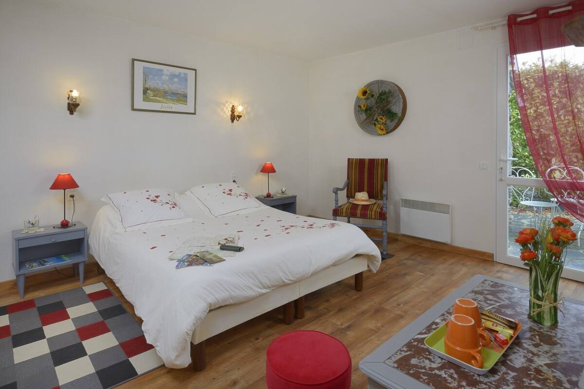 Une chambre à coucher comprenant un lit, des tables de chevet, une chaise, une petite table avec un plateau, une fenêtre avec des rideaux et un mur orné d’un tableau et de quelques appliques murales.