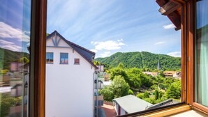 A view from a window overlooking a white building with a sloped roof, surrounded by lush greenery and a mountain in the background. - Hotel Ristorante Piccolo (Thale)
