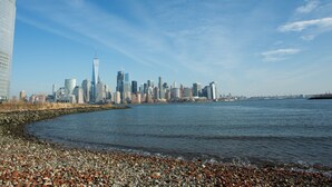 A waterfront cityscape with a prominent skyscraper, a rocky shoreline, and a clear blue sky. - 1 Bedroom Apt Hoboken  - Close to NYC/MetLife - FIFA (Hoboken)