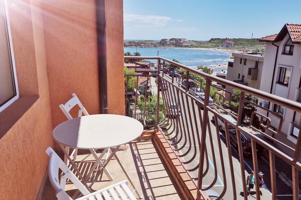 A balcony with a table and chairs, overlooking a beach and a parking area with cars.