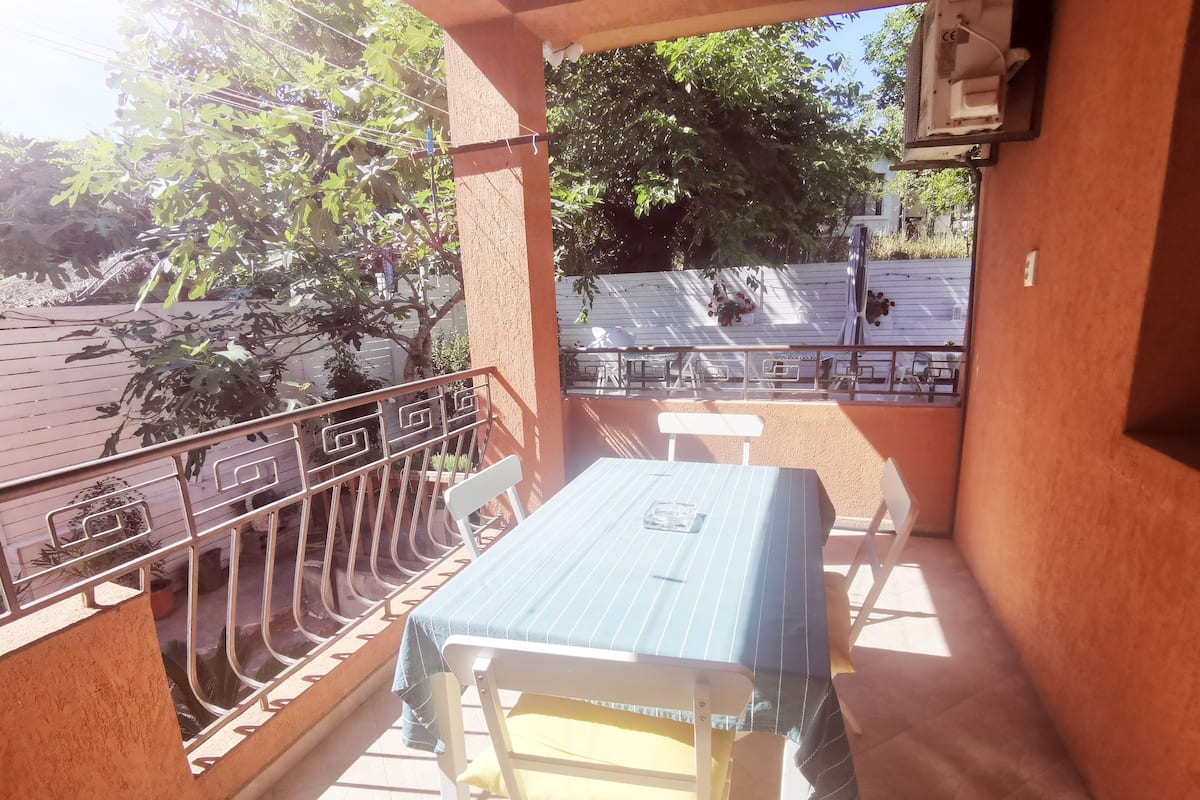 A balcony with a table and chairs, a railing, and a view of trees and a building in the background.