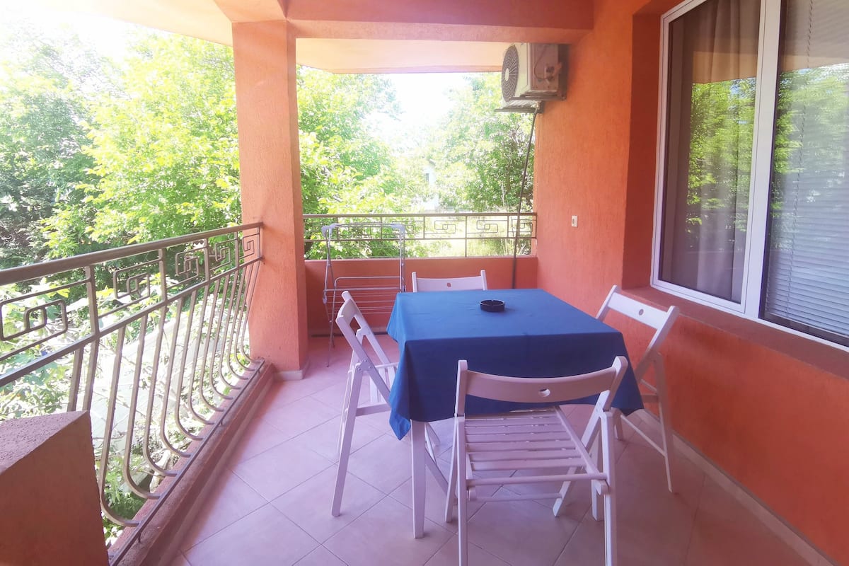 A balcony with a table and chairs, a window with blinds, and a view of trees.