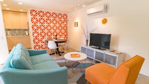 Ein modernes Wohnzimmer mit Salbeigrün- und Orangefarbenen Sofas, einem Esstisch mit Stühlen, einem Fernseher auf einem Schrank und einer Klimaanlage an der Wand.