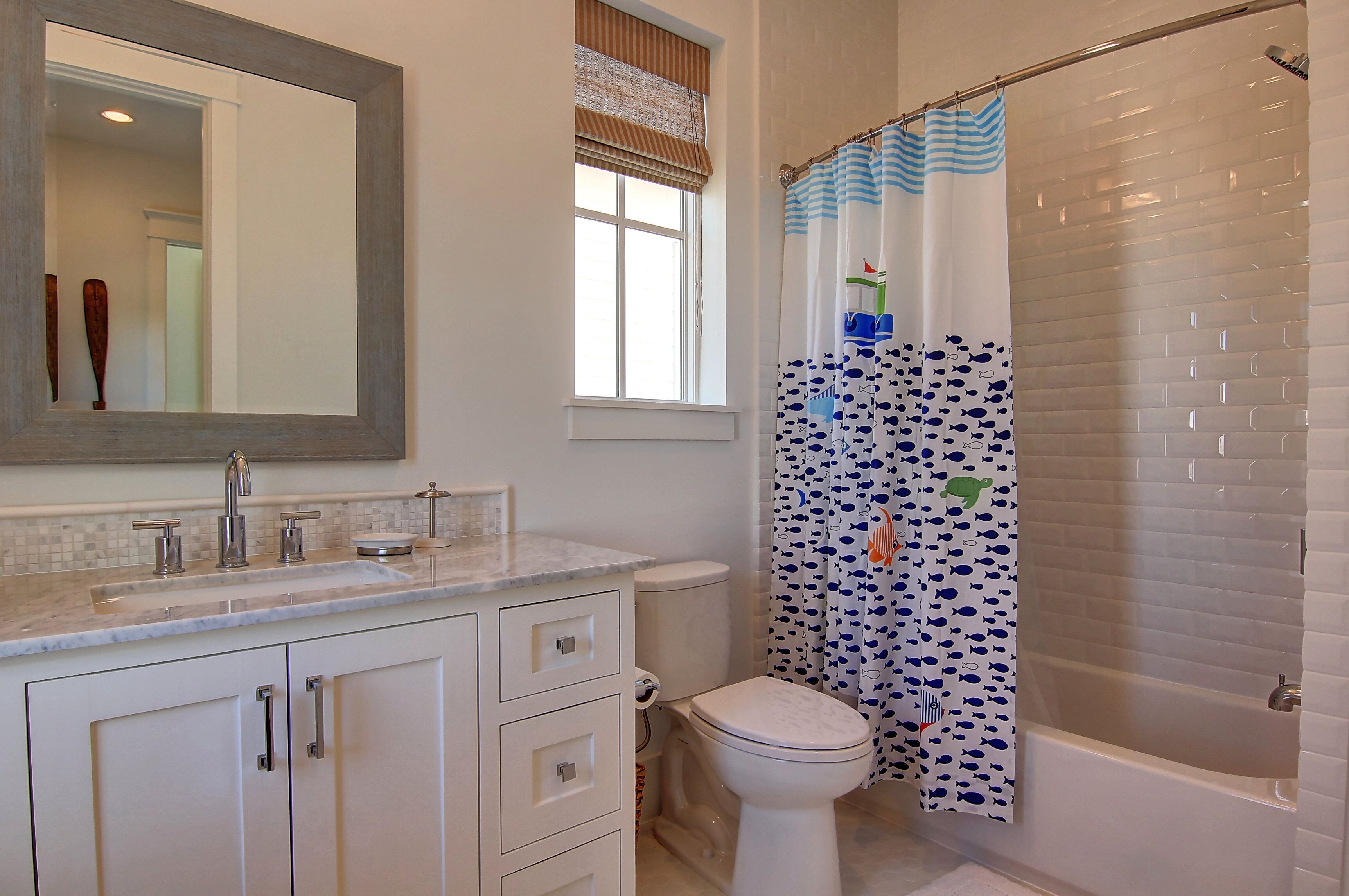 Une salle de bain avec des toilettes blanches, un lavabo avec un miroir au-dessus, un rideau de douche orné d’un motif de poissons et une baignoire.